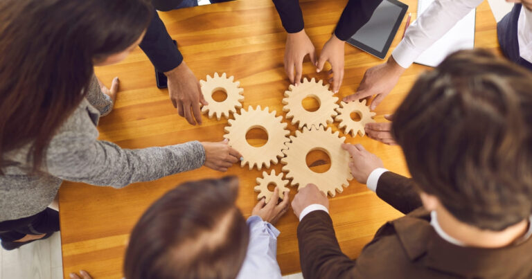 Personas colaborando alrededor de engranajes de madera como símbolo de trabajo en equipo e innovación sin presupuesto.